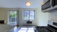 This single-floor unit presents a compact, open-concept layout where the kitchen and living area flow seamlessly together. The kitchen is equipped with a gas range stovetop and a wall-mounted microwave, complemented by granite countertops that add a polished touch to the space. Tile flooring runs throughout the kitchen area, reflecting natural light and contributing to the bright, airy feel of the interior.

The living area receives generous daylight through a double-pane window, which also provides views of surrounding greenery and neighboring buildings. A wall-mounted air conditioning unit sits beneath the window, paired with a baseboard heating system, ensuring year-round climate control. A sliding glass door leads to an outdoor balcony, further enhancing the sense of openness and offering an additional connection to the outdoors.

The space is illuminated by a centrally mounted flush ceiling light, complementing the natural light throughout the day. The white walls and light-toned surfaces throughout the unit create a clean, neutral aesthetic, making the space feel open and well-maintained overall.