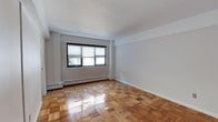 This single-floor space presents a clean, understated aesthetic with a timeless appeal. The standout feature is the rich parquet hardwood flooring, laid in a classic block pattern with a warm honey tone and a polished finish that reflects light beautifully throughout the room. Crisp white walls and a smooth white ceiling create a bright, neutral backdrop that lends the space a sense of openness and versatility. A black-framed window fitted with horizontal blinds allows natural light to filter in, illuminating the interior while offering privacy control. A baseboard heating unit runs along the wall beneath the window, ensuring efficient and consistent warmth throughout the space. The room's simple, rectilinear architecture allows for flexible furniture arrangements, making it highly adaptable to a variety of uses. Clean white baseboards frame the perimeter of the room, adding a subtle finishing touch that ties together the overall design. The combination of warm wood flooring, neutral walls, and natural light gives the space a welcoming and functional ambiance suitable for a range of occupants.
