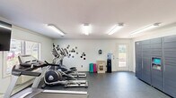 This fitness center in Columbia, SC is a well-organized, single-room exercise facility designed to support a well-rounded workout routine. The space features durable dark flooring throughout, bright overhead fluorescent lighting, and a clean, light-toned interior that creates an open and motivating atmosphere. Large mirrors along one wall visually expand the room while allowing users to monitor their form during exercise. The cardio equipment selection is comprehensive, including multiple treadmills, elliptical machines, and a recumbent stationary bike, all arranged to maximize the available floor area. A cable resistance machine offers strength training options, and exercise mats are available for stretching and floor work. A wall-mounted flat-screen television is positioned for visibility from the cardio equipment. Natural light enters through windows with blinds, offering views of the surrounding outdoor greenery. A water cooler station provides convenient hydration access. The space also includes a bank of personal storage lockers, offering secure storage for users. A restroom is accessible directly within the facility. A door leads to the outdoors, enhancing ventilation and connectivity to the exterior. The overall design is functional and efficient, providing residents with a practical and fully equipped space for daily fitness.