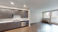 This single-floor unit showcases a clean, contemporary design throughout. The open-concept living and kitchen area is anchored by wood-style laminate flooring that flows seamlessly through the main space, creating a cohesive and inviting atmosphere. Neutral light grey walls and white ceilings enhance the brightness of the interior.

The kitchen is well-appointed with flat-front grey cabinetry extending to the upper walls, offering generous storage space. White countertops provide ample prep area, while stainless steel appliances — including a refrigerator, range with hood vent, and dishwasher — complete the functional and modern aesthetic. Recessed lighting illuminates the kitchen efficiently.

The living area benefits from natural light through a large window and a glass-paneled exterior door fitted with blinds, providing both brightness and privacy. A wall-mounted HVAC unit ensures climate control year-round.

The bathroom features wood-style flooring, a vanity with dark cabinetry, grab bars for accessibility, and a clean, minimalist finish. Additionally, a walk-in closet with dual hanging rods and carpeted flooring provides practical and well-organized storage. The overall ambiance of the unit is modern, functional, and move-in ready.