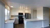 This single-floor space centers around a well-appointed kitchen designed for both functionality and style. The kitchen features crisp white upper and lower cabinetry that provides ample storage, complemented by a mosaic tile backsplash in neutral tones that adds a subtle decorative touch. A suite of appliances includes a black electric range, an over-the-range black microwave, and a stainless steel refrigerator, offering a contemporary contrast to the white cabinetry. A sink with a window-adjacent position sits along the perimeter countertop, enhancing the workspace. The standout feature of the kitchen is a generously sized peninsula with a clean white surface and vertical shiplap-style paneling, providing additional prep space and casual seating potential. Track lighting overhead ensures the workspace is well-illuminated. The flooring throughout is a warm-toned wood laminate, adding warmth and cohesion to the space. A textured ceiling adds a traditional finish. A hallway visible from the kitchen suggests additional rooms beyond, contributing to the overall functional layout of this single-floor space.