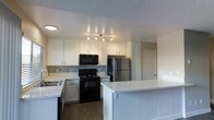 This single-floor space centers around a well-appointed kitchen designed for both functionality and style. The kitchen features crisp white upper and lower cabinetry that provides ample storage, complemented by a mosaic tile backsplash in neutral tones that adds a subtle decorative touch. A suite of appliances includes a black electric range, an over-the-range black microwave, and a stainless steel refrigerator, offering a contemporary contrast to the white cabinetry. A sink with a window-adjacent position sits along the perimeter countertop, enhancing the workspace. The standout feature of the kitchen is a generously sized peninsula with a clean white surface and vertical shiplap-style paneling, providing additional prep space and casual seating potential. Track lighting overhead ensures the workspace is well-illuminated. The flooring throughout is a warm-toned wood laminate, adding warmth and cohesion to the space. A textured ceiling adds a traditional finish. A hallway visible from the kitchen suggests additional rooms beyond, contributing to the overall functional layout of this single-floor space.