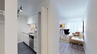 This single-floor residence showcases a thoughtfully designed layout with a modern aesthetic defined by clean lines, neutral tones, and light wood flooring throughout the main living areas. The open-concept design connects the kitchen directly to the living space, creating a seamless flow ideal for everyday living and entertaining.

The kitchen is outfitted with sleek white cabinetry, dark tile flooring, stainless steel appliances including an electric range with over-the-range microwave, a built-in dishwasher, and a undermount sink. White subway tile backsplash adds a crisp, contemporary touch.

The living area is bright and inviting, featuring large floor-to-ceiling windows dressed with vertical blinds that allow generous natural light. A distinct seating nook accommodates a bistro-style table with bar stools alongside a sculptural egg chair, adding a design-forward element to the space.

Two bedrooms provide comfortable retreats, each with light wood flooring or plush carpet, large windows with vertical blinds, and ample wall space. The bathroom is finished in an all-white palette with hexagonal tile flooring, a modern vanity with an undermount sink, and a shower-tub combination with curtain enclosure.