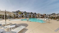 Located at 1110 Mason Pond Pl in Ladson, South Carolina, this outdoor amenity space serves as the centerpiece of a well-appointed residential community. The highlight of the area is a generously sized resort-style swimming pool with a freeform design, its clear turquoise waters set against a broad, light-colored concrete sundeck. The deck is furnished with numerous white lounge chairs and market umbrellas, providing ample space for sunbathing and relaxation. A covered pergola structure anchors one side of the pool area, offering a shaded gathering spot with additional seating. Ornamental landscaping is thoughtfully integrated throughout, featuring palm trees, flowering shrubs, and manicured plantings that lend a coastal, resort-like ambiance to the setting. A covered entryway with decorative hanging flower baskets and classic column detailing provides an inviting transition into the pool area, framed by wrought iron fencing. Vintage-style lamp posts line the walkways, adding charm and functionality. The surrounding multi-story residential buildings feature craftsman-inspired architecture with gabled rooflines, private balconies, and dark-trimmed windows. The overall atmosphere is bright, open, and welcoming, reflecting a community designed for active outdoor living under the sunny South Carolina sky.