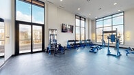 This contemporary fitness center is designed to deliver a premium workout experience within a bright, open, and architecturally striking environment. The space is defined by soaring double-height ceilings and expansive floor-to-ceiling windows framed in sleek black steel, flooding the interior with abundant natural light and offering open views of the surrounding neighborhood. The durable rubberized flooring provides both safety and comfort throughout the training area.

The facility is comprehensively outfitted with a wide array of blue commercial-grade strength equipment, including multiple cable and functional trainer machines, a full dumbbell rack spanning a broad weight range, adjustable benches, a power rack, and various specialty strength stations. Large wall-mounted mirrors along one side of the space enhance the sense of depth while allowing users to monitor their form. A wall-mounted flat-screen television adds an entertainment element to the training environment.

A glass partition wall separates the fitness center from an adjacent lounge or common area, maintaining visual connectivity between the spaces while defining distinct zones. Recessed ceiling lighting complements the natural light, ensuring the space remains well-illuminated at all times. The overall ambiance is clean, modern, and motivating, making this fitness center a standout amenity for its users.