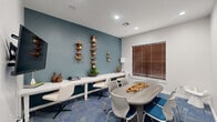 This single-room conference and workspace at 2323 Eldridge Parkway in Houston, Texas, is thoughtfully designed to support both collaborative meetings and individual work. The room is defined by a crisp white ceiling with recessed lighting, paired with a striking teal accent wall that adds depth and visual interest to the otherwise neutral white walls. Blue patterned carpet tiles cover the floor, lending a contemporary, professional character to the space.

At the center of the room sits an oval conference table surrounded by modern mesh-back chairs in white and blue, providing comfortable seating for group gatherings. Along the teal accent wall, a long white workstation counter with additional seating offers a dedicated area for focused individual tasks. Vertical wooden wall-mounted shelving units display small plants and decorative objects, contributing warmth and personality to the workspace.

On the opposing white wall, a large framed world map artwork serves as a bold focal point, accompanied by a wall-mounted flat-screen television, making the space well-suited for presentations. A window dressed with warm wood-toned horizontal blinds allows natural light to filter in. A frosted glass-paneled door provides access to the broader facility, completing this efficient and stylish professional environment.
