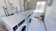 This single-floor laundry facility is a well-equipped, communal space designed with practicality and efficiency in mind. The room features a row of modern front-loading washing machines and dryers, all finished in white, and arranged neatly to maximize the use of available space. The machines are positioned to allow comfortable access, with flat-top surfaces that can double as folding areas. Natural light enters through a window along one wall, brightening the interior and offering a view of the surrounding greenery outside. The walls are painted in a light, neutral tone, contributing to a clean and utilitarian atmosphere. Exposed utility piping is visible overhead, characteristic of a purpose-built laundry space. The flooring is smooth and easy to maintain, suited to the demands of a high-use utility area. A waste bin is conveniently placed near the machines for easy disposal. Overall, this laundry facility provides a straightforward and functional environment, thoughtfully arranged to serve the practical needs of its users efficiently.