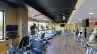 This fitness center offers a comprehensive workout environment spread across a single open floor. The space is divided into distinct functional zones, each thoughtfully arranged to accommodate a variety of exercise needs. The cardio section lines the perimeter near large windows, housing a row of treadmills, elliptical trainers, stationary bikes, and stair climbers—all facing outward to take advantage of natural light and outdoor views of surrounding greenery. The windows contribute to an airy, open atmosphere throughout.

The strength training area occupies the interior of the space, featuring Cybex cable machines and multi-function weight stations positioned for efficient use. A dedicated free weights zone includes a full dumbbell rack and adjustable bench, while stability balls and a BOSU trainer are available for functional fitness exercises. Wall-mounted mirrors extend the visual depth of the room and support form-checking during workouts.

The flooring throughout is a durable hardwood-style surface, providing both a clean aesthetic and practical functionality. The ceiling is finished in a dark matte tone with exposed industrial-style lighting, creating a contrast against the warm yellow walls. Wall-mounted flat-screen televisions are positioned throughout, enhancing the workout experience. The overall ambiance is energetic yet functional.