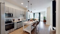 Located at 7488 Universal Blvd in Orlando, FL, this single-floor residence offers a thoughtfully designed open-concept layout with contemporary finishes and a cohesive modern aesthetic. The kitchen is a standout feature, equipped with gray shaker-style cabinetry, white quartz countertops, a classic subway tile backsplash, and a full suite of stainless steel appliances including a refrigerator, range, microwave, and dishwasher. A movable white kitchen island with bar seating and dual pendant lights serves as a functional centerpiece, seamlessly connecting the kitchen to the living area. The living room is bright and welcoming, with large windows dressed in teal drapes that allow generous natural light and views of the surrounding outdoor environment. Wood-look flooring runs throughout the main living spaces, adding warmth and continuity. The bedroom features plush carpeting, a bold teal accent wall, large windows, and ample room for generous furnishings. The primary bathroom is well-appointed with a dual-sink vanity, gray cabinetry, a wide illuminated mirror, and direct access to a walk-in closet. An additional nook provides a dedicated workspace. The overall ambiance is modern, clean, and inviting, with a carefully curated color palette of blues, grays, and warm wood tones.