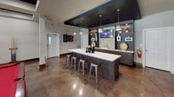 This sophisticated single-floor entertainment and lounge space is thoughtfully designed for social gatherings and relaxation. The centerpiece of the space is a sleek wet bar area, complete with a long white countertop, dark wood cabinetry, open display shelving with decorative accents, a wall-mounted TV, and stylish pendant lighting suspended from a bold dark ceiling tray. Industrial-style metal barstools line the island, adding an urban edge to the design.

The open main area features polished concrete floors that reflect light beautifully, structural columns with contrasting dark bases, and a striking coffered ceiling with dark inset panels that lend architectural depth to the room. This zone accommodates both a billiards table with a classic red felt surface and a comfortable lounge seating area with a sofa, armchairs, and a TV — perfect for entertaining or unwinding.

A separate, more intimate TV lounge room offers a quieter retreat, furnished with bold black-and-white patterned armchairs accented by red throw pillows, wall-mounted artwork, and natural light streaming through multiple windows. The warm, neutral walls and rich dark wood baseboards tie the entire space together, creating a cohesive, modern-yet-inviting atmosphere well suited for community or private entertainment use.