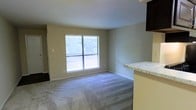 This single-floor residential space offers a clean, functional layout designed for comfortable everyday living. The main living area is carpeted in a soft, neutral-toned plush carpet that adds warmth underfoot and contributes to a quiet, inviting atmosphere. A large window floods the room with natural light while framing a view of lush greenery outside, creating a pleasant connection to the outdoors. The neutral beige wall tones throughout provide a versatile backdrop that complements a variety of décor styles. Adjacent to the living area, a partial kitchen counter features a light granite-style surface with dark cabinetry above, hinting at a well-appointed kitchen space. The kitchen and living areas flow together in an open-concept arrangement, making the space feel connected and easy to navigate. The layout also appears to include additional rooms accessible via a hallway, providing separation between living and private spaces. Overall, the unit presents a straightforward, move-in-ready environment with practical finishes, ample natural light, and a calm, neutral aesthetic throughout.