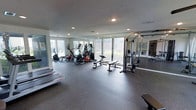 This fitness center is designed to accommodate a comprehensive workout experience within a bright, open environment. Large floor-to-ceiling windows line the perimeter, allowing abundant natural light to fill the room while offering views of the surrounding greenery, creating an energizing atmosphere for exercise. The space features durable rubber flooring throughout, providing both safety and comfort during workouts. Cardio equipment, including elliptical trainers and stationary bikes, is positioned along the windowed wall, taking full advantage of the natural light and outdoor views. A multi-function cable machine and a weight bench are centrally located, offering versatile strength training options. Recessed lighting in the textured ceiling supplements natural light, ensuring the space remains well-lit at all times. Full-length mirrors are mounted along one wall, enhancing the sense of space while allowing users to monitor their form. Additional equipment, including what appears to be rowing machines, is positioned along the opposite wall. The overall layout is thoughtfully organized to maximize usable floor space, promoting ease of movement between equipment stations.