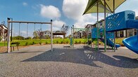 Located at 50 Fenway Place in Concord, North Carolina, this outdoor community amenity space offers a well-appointed playground and recreational area designed to serve residents of the surrounding multi-story residential buildings. The playground is the centerpiece of the space, featuring a vibrant blue and green play structure complete with a spiral slide, climbing elements, and overhead monkey bars. A shade canopy covers a portion of the play area, providing relief from the sun while the impact-absorbing rubberized gravel surface underfoot ensures a safer play environment. Standalone swing posts and additional fitness-style equipment extend the recreational offerings beyond the main structure. The grounds surrounding the playground are thoughtfully landscaped with manicured mulched beds, ornamental shrubs, and lush open lawns that invite outdoor activity. A covered pavilion is visible in the background, adding a gathering space for residents. The surrounding residential buildings feature a contemporary craftsman-style design with stone and board-and-batten siding, balconies, and large windows. Ample surface parking, wide sidewalks, and tall light poles ensure accessibility and safety throughout. The space benefits from an open sky setting with abundant natural light and expansive views of the community grounds.