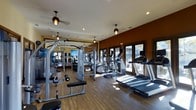 This fitness center offers a comprehensive workout environment designed to accommodate a wide range of exercise needs. The space features warm hardwood-style flooring throughout, which pairs with rich, earth-toned accent walls to create an inviting yet energizing atmosphere. Recessed lighting and stylish pendant fans are mounted along the ceiling, ensuring the space is well-lit and well-ventilated during workouts.

The cardio section is equipped with multiple treadmills and elliptical machines, each fitted with individual display screens, and positioned along a wall of floor-to-ceiling glass doors that allow natural light to pour in and provide views of the outdoor surroundings. The strength training area features a variety of cable and weight machines, free weights, and benches, offering options for both resistance and functional training.

Full-length mirrors line one wall, visually expanding the space and allowing users to monitor their form. The thoughtful layout provides ample room to move between equipment without feeling crowded. The combination of natural light, warm finishes, and modern equipment makes this fitness center a well-rounded and pleasant facility for daily exercise.