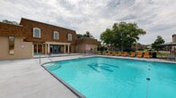 This outdoor aquatic facility centers around a generously sized rectangular swimming pool with crystal-clear turquoise water, complete with lane dividers and pool access ladders at multiple entry points. The pool deck is paved in smooth concrete and lined on all sides with bright orange lounge chairs, offering ample space for sunbathing and relaxation. A separate hot tub is situated adjacent to the main pool, providing a dedicated area for hydrotherapy and leisure. A covered umbrella station with additional seating offers a shaded retreat along the deck perimeter.

The facility is anchored by a two-story brick clubhouse building featuring arched windows and a covered pergola entrance with blue double doors, lending a classic architectural character to the space. Adjacent to the clubhouse is a smaller utility building with a shingle-style roof, housing restroom and changing facilities accessible directly from the pool deck. The entire area is enclosed by a black iron perimeter fence, ensuring privacy and security, while mature trees beyond the fence provide a natural green backdrop. The open-air setting allows for abundant natural light throughout the day, creating an inviting atmosphere well-suited for community recreation and outdoor gatherings.