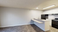 This efficiently designed single-floor unit offers a thoughtful layout comprising a living area, kitchen, and bathroom. The living room features neutral-toned walls, clean white baseboards, and wood-look laminate flooring that runs throughout the space, lending a modern and cohesive feel. A window fitted with horizontal blinds allows natural light to brighten the room. The textured ceiling adds a classic residential touch.

The kitchen is well-appointed and functional, featuring crisp white cabinetry with ample upper and lower storage, light-toned countertops, and a suite of black appliances including a full-size refrigerator, an electric range, and a built-in range hood. A peninsula counter with an integrated sink extends into the living area, creating an open, connected feel between the kitchen and living space. Overhead fluorescent lighting ensures the kitchen is well-lit.

The bathroom is clean and practical, offering a single-sink vanity with white cabinetry and a speckled countertop, a large wall mirror, a combined tub and shower unit, and a wall-mounted grab bar for added accessibility. Wood-look flooring continues into the bathroom, maintaining design consistency throughout.

Overall, the unit presents a straightforward, move-in-ready aesthetic with contemporary finishes and efficient use of its compact footprint.