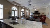 This single-floor space showcases a grand, open-concept layout that seamlessly connects a kitchen area to a spacious lounge and living area. The kitchen features a thick marble-veined granite countertop with an undermount sink and a high-arc faucet, offering both style and functionality. Overhead, geometric black metal pendant chandeliers hang as striking focal points, adding a modern industrial contrast to the otherwise classic interior.

The living area is anchored by a stone-clad fireplace flanked by comfortable upholstered seating, creating a warm and inviting atmosphere. Tall arched windows with decorative transoms line the walls, flooding the space with abundant natural light and offering views of the surrounding greenery. The soaring ceilings, accentuated by clean crown molding and column details, lend a sense of grandeur throughout.

Neutral warm tones on the walls complement the rich furnishings, including plush sofas and layered rugs that define distinct seating zones. A wall-mounted television is positioned above the fireplace, adding a practical entertainment element to the elegant setting. The overall ambiance is refined yet comfortable.