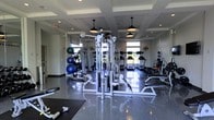 This fitness center is thoughtfully designed to support a full range of workout routines in a clean, modern environment. The centerpiece of the space is a multi-station cable and weight machine, offering a wide variety of strength training options for users of all fitness levels. Surrounding it, a selection of free weights, weight plates, and stability balls are neatly arranged along mirrored walls, which enhance the sense of openness and allow users to monitor their form during exercise. Toward the rear of the room, a row of treadmills and additional cardio equipment lines the space beneath large windows that invite natural light and provide outdoor views, creating an energizing atmosphere. The flooring is a polished, dark surface that is both durable and easy to maintain. Overhead, a coffered ceiling with recessed lighting and stylish industrial-style pendant lights adds an architectural touch that elevates the overall aesthetic. Ventilation fans are strategically positioned to ensure comfort during intense workouts. The layout is open and well-organized, allowing for easy movement between equipment zones while maintaining a safe and functional workout environment.