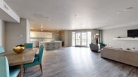 This single-floor community space offers a generously sized, open-plan layout that seamlessly integrates a kitchen, dining area, and living room into one cohesive environment. The wood-look plank flooring runs throughout, tying each zone together while complementing the neutral gray and white color palette accented by teal furnishings.

The kitchen is well-appointed with white shaker cabinetry, stainless steel appliances including a refrigerator and dishwasher, a light blue subway tile backsplash, and crisp white countertops. A central island with bar-height seating provides additional prep space and casual dining options. Track and pendant lighting illuminate the workspace efficiently.

Adjacent to the kitchen, a dedicated dining area accommodates a full-sized table with upholstered teal chairs, making it suitable for group gatherings. Coastal-themed artwork adorns the walls, adding a relaxed, inviting atmosphere.

The living area features a large sectional sofa, a wall-mounted flat-screen television on a floating shelf, and a glass-top coffee table set atop a teal area rug. Glass-paneled interior doors and a set of full-height French doors opening to an outdoor patio flood the space with natural light and provide views of the exterior. The textured ceiling and recessed lighting complete the polished, functional design of this versatile shared space.