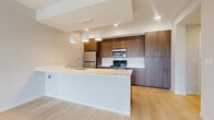 This contemporary single-floor residence offers a thoughtfully designed open-concept layout with modern finishes throughout. The kitchen serves as a central highlight, featuring rich walnut-toned cabinetry extending to the ceiling, a generous white quartz waterfall island with an integrated sink and gold-toned faucet, stainless steel appliances including a refrigerator, range, and built-in microwave, and a trio of pendant lights suspended above the island. Light wood-look flooring flows throughout the main living areas, complementing the clean, neutral palette. The spacious living area is bathed in natural light through large windows and a sliding glass door that opens to a private balcony overlooking surrounding greenery and open landscape views. Bedrooms feature soft carpeting and oversized windows framing tranquil outdoor scenery. The bathroom is finished with a wood-grain vanity, white quartz countertop, and brushed gold hardware, maintaining the cohesive modern aesthetic. A dedicated laundry closet with a stacked full-size washer and dryer adds practical convenience. Recessed lighting and tray ceiling details in the kitchen further elevate the overall design, creating a polished and comfortable living environment.
