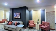 This single-floor space presents a contemporary lounge area designed with a refined, modern aesthetic. The walls are finished in a deep charcoal tone, creating a sophisticated and intimate atmosphere that contrasts effectively with the recessed ceiling lighting overhead. A neutral-toned sofa accented with coral and dark throw pillows anchors one side of the room, paired with a sleek, minimalist side table. Across the space, two tufted coral leather armchairs flank a light-colored marble-style side table, offering a cohesive and inviting seating arrangement. The standout feature of the room is an illuminated textured wall panel in warm golden tones, lending a dramatic and visually striking element to the corner. A vibrant piece of wall art displaying an abstract cityscape adds a pop of color against the dark backdrop. The patterned carpet introduces subtle visual texture underfoot, grounding the overall design. The combination of warm accent lighting, bold wall tones, and carefully curated furnishings gives this space a polished, professional ambiance well-suited for a reception, hospitality, or lounge setting.