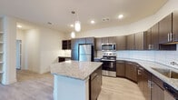 This well-appointed single-floor unit showcases a thoughtfully designed open-concept layout with light wood-style flooring that flows seamlessly throughout the space. The kitchen serves as the heart of the home, featuring a large central island topped with granite countertops and illuminated by elegant pendant lighting. Dark espresso cabinetry lines the walls, providing generous storage, while a full suite of stainless steel appliances—including a refrigerator, range, microwave, and dishwasher—adds a sleek, modern touch. A classic white subway tile backsplash ties the design together, and a built-in desk area offers a convenient workspace. The open layout connects the kitchen to the living area, which benefits from natural light and includes built-in shelving for added functionality. The bedrooms are bright and airy, each featuring light wood-style flooring, windows with blinds, and ceiling fans for comfort. At least one bedroom includes access to a walk-in closet with wire shelving. The bathrooms are finished with granite-topped dark wood vanities, framed mirrors, and shower/tub combinations, maintaining the cohesive modern aesthetic found throughout the unit. The overall ambiance is clean, contemporary, and move-in ready.