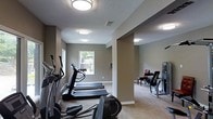 This fitness center in Arlington, Texas, is a well-appointed, single-floor facility designed to support a complete workout routine. The space is thoughtfully laid out to accommodate both cardio and strength training activities, with dedicated zones for each type of exercise. The cardio area features multiple treadmills and an elliptical machine, arranged in a row along neutral-toned walls that create a calm, focused atmosphere. A window at the far end of the cardio zone allows natural light to filter in, adding a welcoming brightness to the space. The flooring throughout is a durable, textured carpet well-suited for a fitness environment. Flush-mounted ceiling lights provide consistent, even illumination across the entire facility. An adjacent area houses strength training equipment, including a weight bench and cable machine, separated from the cardio zone by a structural column that helps define each workout area without fully enclosing the space. The textured ceiling and clean white baseboards contribute to a tidy, maintained appearance. The overall ambiance is practical and inviting, offering a comfortable environment for residents or members to pursue their fitness goals in a well-equipped, organized setting.
