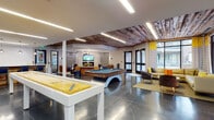 This single-floor amenity lounge is thoughtfully designed to offer a dynamic, multi-functional social environment. The space is divided into several distinct zones that flow seamlessly into one another across polished concrete tile flooring. A prominent game area anchors the central portion of the room, featuring a full-size billiards table and a shuffleboard table, complemented by high-top seating and a large wall-mounted television along a stacked stone accent wall.

A dedicated lounge zone showcases an electric fireplace set into a stacked stone surround, topped by a mounted flat-screen TV. Upholstered seating panels flank the fireplace wall, while a leather sofa, live-edge wood coffee table, and a sculptural multi-bulb chandelier create an inviting, refined atmosphere.

Near the floor-to-ceiling windows and glass door leading to an outdoor area, a casual seating nook features a sectional sofa with bold yellow accent pillows, bathed in natural light filtered through patterned curtains. Reclaimed wood ceiling panels add warmth and texture to portions of the space, contrasting with the clean modern finishes elsewhere.

A separate kitchenette area offers dark cabinetry with white countertops, stone-clad walls, pendant lighting, and a glazed door connecting to an adjacent corridor, providing practical support for social gatherings.