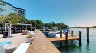 This exceptional waterfront property offers direct access to open water via a well-appointed private dock, making it a premier destination for boating and coastal living. The expansive wooden dock stretches along the waterline, featuring multiple boat slips equipped with mooring posts capable of accommodating several vessels simultaneously. The dock's warm hardwood decking is both functional and visually appealing, creating a seamless transition between the property and the water.

Adjacent to the dock, a relaxed lounge area provides comfortable seating with sun loungers and decorative planters filled with tropical greenery, offering a tranquil space to enjoy the waterfront views. A pergola structure provides welcome shade, enhancing the outdoor living experience.

The surrounding landscape is rich with mature palm trees and lush tropical vegetation, framing the property with natural beauty and privacy. The calm, clear waters reflect the vibrant Florida-style environment, ideal for boating, fishing, or simply soaking in the scenery. A multi-story building is visible on the property, suggesting additional accommodations or amenities. Under clear blue skies, this property delivers an unmatched combination of practical marina-style amenities and a relaxed, resort-like ambiance.
