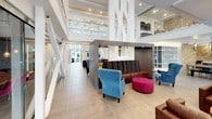 This thoughtfully designed amenity space showcases a striking blend of contemporary architecture and inviting interiors across a single, well-appointed floor. The centerpiece is a dramatic double-height lobby flooded with natural light through expansive windows, featuring sculptural white columns, a glass-railed mezzanine overlook, and large-format porcelain tile flooring. Eclectic seating arrangements — including rich leather sofas, vibrant blue wingback chairs, and a bold magenta ottoman — create a welcoming, social atmosphere throughout the open common area.

A dedicated fireplace lounge offers a more intimate setting, anchored by a floor-to-ceiling stacked stone surround with a linear gas fireplace, comfortable leather club chairs, and direct access to an outdoor patio. The kitchen area is outfitted with sleek dark cabinetry, white countertops, a stainless steel Bosch wall oven, and a textured stacked stone accent wall with open shelving.

Private workspaces include a bright office with large windows, a clean-lined white desk, and an adjacent glass-enclosed conference room with its own distinct furnishings. Throughout, thoughtful details such as bold geometric wallcovering in the restroom corridor, dual water fountains, and abundant recessed lighting contribute to the polished, modern character of this versatile community space.