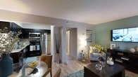 This single-floor residence showcases an open-concept design that connects the kitchen, dining, and living areas into one cohesive, flowing space. The kitchen is equipped with stainless steel appliances, including a refrigerator, microwave, and range, complemented by dark cabinetry that provides a striking contrast against the light-toned walls. Light wood-style flooring runs throughout, lending warmth and continuity across the space.

The dining area transitions naturally into the living room, which features a soft sage-green accent wall that adds a calming, contemporary touch. A plush armchair, tasteful décor, and ambient floor lighting contribute to the inviting atmosphere. A textured area rug anchors the seating area, adding dimension underfoot.

A hallway visible from the main living area leads to additional rooms, suggesting a well-organized and functional floor plan. The consistent neutral color palette of warm grays and greens, combined with carefully placed decorative elements such as tall branch arrangements and floral accents, gives the space an aesthetically curated and welcoming character throughout.