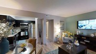 This single-floor residence showcases an open-concept design that connects the kitchen, dining, and living areas into one cohesive, flowing space. The kitchen is equipped with stainless steel appliances, including a refrigerator, microwave, and range, complemented by dark cabinetry that provides a striking contrast against the light-toned walls. Light wood-style flooring runs throughout, lending warmth and continuity across the space.

The dining area transitions naturally into the living room, which features a soft sage-green accent wall that adds a calming, contemporary touch. A plush armchair, tasteful décor, and ambient floor lighting contribute to the inviting atmosphere. A textured area rug anchors the seating area, adding dimension underfoot.

A hallway visible from the main living area leads to additional rooms, suggesting a well-organized and functional floor plan. The consistent neutral color palette of warm grays and greens, combined with carefully placed decorative elements such as tall branch arrangements and floral accents, gives the space an aesthetically curated and welcoming character throughout.