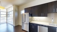 This contemporary single-floor space is designed with a clean, modern aesthetic that emphasizes both functionality and style. The kitchen serves as a standout feature, showcasing rich dark cabinetry that extends to the ceiling for maximum storage capacity. Stainless steel appliances, including a French door refrigerator and dishwasher, complement the modern design palette. The countertops are finished in granite, offering both durability and an elegant visual contrast against the dark cabinetry. A classic subway tile backsplash with under-cabinet lighting adds warmth and practicality to the cooking area. The flooring throughout is a dark hardwood, providing a cohesive and sophisticated foundation that ties the space together. Light-toned walls keep the interior feeling bright and open, while strategically placed windows allow natural light to filter in, enhancing the overall ambiance. The open layout creates a seamless flow between the kitchen and adjoining areas, making the space feel inviting and well-connected. The combination of high-quality finishes, thoughtful design details, and a neutral color palette gives this space a timeless, move-in-ready appeal suited for modern living.
