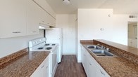 This single-floor space is designed with functionality and clean aesthetics in mind. The kitchen serves as a central highlight, laid out in a classic galley configuration that maximizes efficiency within a narrow footprint. White cabinetry lines both sides of the kitchen, offering ample storage space from floor to ceiling, while dark granite-style laminate countertops provide a warm contrast to the bright, white finishes. A white electric range with coil burners and a matching full-size refrigerator are seamlessly integrated into the layout, complementing the overall cohesive color palette. The double-basin stainless steel sink sits beneath upper cabinets on the opposing counter, adding a practical touch. Rich, dark hardwood-style flooring runs the length of the kitchen, grounding the space with warmth and visual depth. The walls are finished in a clean, neutral tone, contributing to the bright and open feel of the room. A range hood above the stove ensures proper ventilation, and recessed ceiling lighting keeps the space well-lit. The corridor-style layout leads naturally to adjacent areas of the home, maintaining a smooth and connected flow throughout the single-level floor plan.