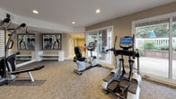 Located at 2502 Manor Dr in Fredericksburg, Virginia, this well-appointed fitness center offers a comprehensive workout environment designed for both cardio and strength training. The space is organized across an open, single-level layout with neutral-toned walls, recessed lighting, and plush carpeted flooring throughout.

The cardio zone is a standout feature, lined with floor-to-ceiling sliding glass doors and large windows that flood the area with natural light and provide views of the surrounding outdoor patio and landscaping. This section houses multiple treadmills and an elliptical trainer, all equipped with modern digital displays. A recumbent stationary bike and rowing machine add further cardio variety.

A dedicated strength training area features a multi-station cable and weight machine alongside free weights and exercise balls. Full-length mirrored walls line this section, creating a sense of expanded space while allowing users to monitor their form. Built-in white bench seating provides a functional rest area, and motivational fitness artwork adorns the walls, enhancing the athletic atmosphere.

A secondary mirrored room offers additional space for stretching or group exercise with stability balls and accessories. Sliding glass doors throughout provide convenient direct access to the outdoor patio area, seamlessly blending the indoor fitness environment with the surrounding exterior.