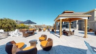This expansive outdoor patio and courtyard space is thoughtfully designed for both relaxation and social gatherings. The ground-level area features large-format stone pavers throughout, creating a clean and cohesive aesthetic across the entire space.

Multiple distinct seating zones are arranged across the patio, including stylish wicker lounge chairs with vibrant orange cushions, modular sectional sofas, and round dining tables shaded by large patio umbrellas. A central focal point is a beautifully crafted circular fire pit, constructed with stacked stone and a stainless steel surround, encircled by classic white Adirondack chairs — ideal for gathering on cooler evenings.

A covered pergola with warm wood-toned posts and a pitched roof anchors one side of the patio, providing a sheltered outdoor area with additional seating. The adjacent building features a horizontal lap siding exterior in neutral tones, with large multi-pane windows and glass doors that connect the indoor and outdoor spaces seamlessly.

The patio is enclosed by a perimeter railing, beyond which expansive views of the surrounding neighborhood, mature trees, and open sky are visible. Sun loungers and additional seating are positioned to take full advantage of natural light and the scenic surroundings. The overall ambiance is open, bright, and welcoming, offering a well-appointed outdoor retreat.