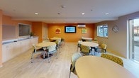 Located at 4051 S 144th St in Tukwila, Washington, this single-floor community gathering space is designed to serve as a functional and inviting dining and lounge area. The space is divided into two distinct zones: a vibrant dining area and a fully equipped kitchen, both thoughtfully appointed for group use.

The dining area is characterized by warm orange accent walls adorned with colorful fruit-themed artwork, creating an energetic and cheerful atmosphere. Light wood-tone flooring runs throughout, contributing to a bright, clean aesthetic. Multiple round tables with patterned upholstered chairs provide ample seating for a variety of group sizes. A wall-mounted flat-screen television anchors one end of the room, while a wall-mounted mini-split HVAC unit ensures year-round comfort. Windows along the perimeter bring in natural light, and a sliding glass door opens directly to an outdoor patio area, extending the usable space.

The adjacent kitchen is well-equipped with full-size black appliances, including a refrigerator, gas range with overhead ventilation, and a coffee maker. White upper cabinetry provides generous storage, and a long counter with an integrated sink doubles as a bar-style seating area. Recessed lighting throughout both areas ensures consistent, practical illumination.