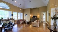 This single-story space is characterized by its striking vaulted ceilings, which lend an airy, open quality to the interior. The walls are finished in warm earth tones, contributing to an inviting and comfortable ambiance throughout. Polished concrete floors run across the main living area, offering both durability and a modern aesthetic touch that contrasts pleasingly with the traditional furnishings.

The main seating area is anchored by two upholstered sofas arranged around a wooden coffee table, providing a relaxed and functional lounge setting. A dark wood media console positioned along one wall houses a television and is flanked by wall-mounted light fixtures that add warm, ambient lighting. Decorative greenery near the console introduces a natural element to the space.

Large windows along one wall allow natural light to filter in, offering views of the surrounding outdoor greenery and brightening the interior considerably. Recessed and track lighting in the ceiling supplement natural light as needed. A French door with glass panels on the right side of the room connects to an adjacent area, adding architectural interest and allowing light to pass through seamlessly.