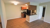 This single-floor residence offers a well-organized open-concept layout that seamlessly connects the kitchen and living areas. The kitchen is a standout feature, equipped with black appliances including a gas range and refrigerator, rich medium-toned wood cabinetry, and striking dark countertops that provide a bold contrast against the light-colored walls. A peninsula with a built-in sink and matching dark countertop defines the boundary between the kitchen and the living area, making it an ideal space for casual dining or entertaining. An overhead range hood anchors the cooking zone with a modern, structured presence. The flooring throughout is warm hardwood-style planking that adds continuity and a sense of warmth to the space. Walls are painted in a soft, neutral tone that enhances the brightness of the room, complemented by flush ceiling lighting that provides even illumination. The layout is efficient and functional, making excellent use of the available space while maintaining an open, airy feel. A sliding door visible along one wall hints at additional access to an exterior area, adding to the overall connectivity of the space.