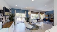 This single-story clubhouse offers a well-appointed communal space designed for comfort and social interaction. The main lounge area is filled with an abundance of natural light, courtesy of expansive floor-to-ceiling windows and sliding glass doors that frame lush, tree-lined outdoor views. The interior is finished with light hardwood-style flooring that enhances the open, airy feel of the space. A striking blue accent wall adds a bold pop of color, complementing the clean white ceiling adorned with recessed lighting and a contemporary pendant lamp. The seating area is thoughtfully arranged with sleek white lounge chairs and a minimalist coffee table, all set atop a patterned area rug. Elegant white drip curtains frame the large windows, while roller shades provide adjustable privacy. Potted tropical plants are strategically placed throughout, bringing a touch of nature indoors. A secondary workspace or activity area is visible toward the rear, offering additional functionality. The overall ambiance is modern, clean, and inviting, making this clubhouse an ideal gathering space for relaxation and community engagement.