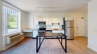 Located at 2015 Lyndale Ave S in Minneapolis, Minnesota, this single-floor unit offers a clean, contemporary design with an efficient and functional layout. The open-concept kitchen and living area serves as the heart of the space, featuring light hardwood-style flooring, white cabinetry, stainless steel appliances—including a refrigerator, range, microwave, and dishwasher—and a built-in countertop workspace. Track lighting runs along the ceiling, providing ample illumination throughout. Large floor-to-ceiling windows flood the living area with natural light and offer views of the surrounding neighborhood, creating a bright and welcoming atmosphere. The living area is generously proportioned, providing flexible space for a variety of furniture arrangements. The bathroom is finished with a white vanity with a light countertop, a wall-mounted mirror, and wood-tone flooring, maintaining the unit's clean aesthetic. A dedicated walk-in closet offers substantial storage with wire shelving along multiple walls and carpeted flooring. The overall design emphasizes simplicity and practicality, with neutral tones and modern finishes throughout that create a cohesive and move-in-ready living environment.