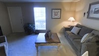 This single-floor space centers around a comfortable living room that balances functionality with a relaxed, homey aesthetic. The room features wall-to-wall carpeting in a neutral tone, contributing to a soft and inviting atmosphere underfoot. A large window along one wall allows natural light to filter into the space, brightening the interior and offering a view of greenery outside. The walls are finished in a warm beige tone that complements the overall palette of the room. A traditional-style coffee table with ornate legs serves as the focal point of the seating area, topped with decorative ceramic vessels that add a touch of character. A plush gray sofa positioned nearby is accented with a large throw pillow, providing a comfortable place to relax. A side table with a classic lamp casts a warm, ambient glow in the corner. Framed artwork above the sofa area adds a decorative touch to the walls. A potted plant near the window introduces a natural element to the space. The overall ambiance is calm and understated, well-suited for everyday living.