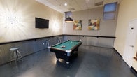 This single-floor game room is designed with recreation and socializing in mind. The centerpiece is a full-sized pool table with a green felt surface, positioned beneath a sleek, dome-shaped pendant light that provides focused illumination. The polished concrete flooring adds an industrial character to the space, complementing the corrugated metal wainscoting that lines the lower portion of the walls — a distinctive architectural detail that gives the room a contemporary, urban feel. A wall-mounted flat-screen television is positioned on one wall, ideal for watching sports or entertainment during gatherings. Decorative starburst wall art and two colorful abstract canvas paintings add visual interest and a modern aesthetic. Bar-height stools are placed along the walls, offering casual seating for spectators. A drop ceiling with recessed and pendant lighting ensures the room is well-lit throughout. The overall ambiance is energetic yet comfortable, making it a well-equipped communal space suited for leisure, entertainment, and informal social gatherings.