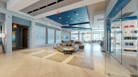 This expansive single-floor amenity space is defined by its contemporary coastal aesthetic, characterized by bold teal ceiling accents, recessed lighting, and large-format beige tile flooring throughout. The open lobby serves as the centerpiece, featuring a dramatic coffered ceiling with vivid blue insets, backlit textured wall panels, and generously spaced lounge seating arranged around sleek coffee tables. Natural light pours in through floor-to-ceiling windows and glass entryways, enhancing the airy, open atmosphere.

A dedicated leasing or reception area includes multiple workstations positioned near bright, curtain-framed windows, with a separate consultation nook enclosed by glass partitions. Adjacent to the main lounge, a stylish bar area offers counter seating with modern black barstools, pendant lighting, and a bold teal accent wall with backlit details.

A glass-enclosed library and conference room showcases illuminated built-in bookshelves alongside a long communal table with seating. The game room features a pool table beneath a statement pendant light, flanked by cafe-style seating and floor-to-ceiling windows offering views of the surrounding exterior. Throughout, curated artwork and cohesive furnishings reinforce the polished, resort-inspired character of the space.