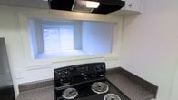 The kitchen in this single-floor space is designed with functionality in mind. It features a black electric coil-burner range as a central cooking appliance, complemented by a sleek overhead exhaust hood that efficiently ventilates the cooking area. The countertops display a speckled granite-style finish, adding a touch of texture and durability to the workspace. A recessed nook above the range, framed in white, provides additional storage or display space and houses a window dressed with vertical blinds that allows natural light to filter into the kitchen, brightening the area. The white cabinetry visible above contributes to a clean, classic aesthetic throughout. The overall design is practical and straightforward, offering the essential elements needed for everyday cooking and meal preparation.