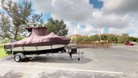 This single-level outdoor space functions as a dedicated storage and parking area, as evidenced by the presence of a trailered boat securely covered with a fitted dark brown cover. The paved surface features clearly marked parking stall lines, providing an organized layout suitable for oversized vehicles and watercraft. The lot is open-air, benefiting from natural light throughout the day, with a backdrop of mature trees lining the perimeter and a residential complex visible beyond a chain-link fence in the background. The surrounding landscape gives the area a suburban Florida character, consistent with communities near waterways where boat storage is a practical necessity. Additional images reveal the exterior of what appears to be a large enclosed trailer with a white paneled sidewall constructed with riveted seams, suggesting durable utility construction. The interior views, though dimly lit, indicate an enclosed storage or cargo space with minimal visibility into its contents. Overall, the space is practical and utilitarian in nature, designed primarily to accommodate the storage and transport of large recreational or commercial equipment, with outdoor accessibility and proximity to a well-established residential neighborhood.