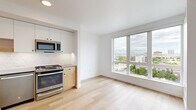 This thoughtfully designed single-floor unit makes efficient use of its compact footprint, combining a clean modern aesthetic with practical functionality throughout. Light hardwood flooring extends across the living area and kitchen, lending warmth and continuity to the open layout.

The galley-style kitchen is well-appointed with stainless steel appliances, including a gas range, dishwasher, microwave, and a full-size refrigerator. Natural wood lower cabinetry is paired with crisp white upper cabinets and a geometric tile backsplash, creating a contemporary two-tone look. A stainless steel sink and ample counter space complete the kitchen. A stacked washer and dryer unit is conveniently integrated near the kitchen corridor.

The living area benefits from an oversized picture window that fills the space with natural light and frames elevated views of the surrounding cityscape, including notable architectural landmarks on the horizon. The bright white walls and clean lines of the room enhance the sense of openness.

The bathroom features a full-size soaking tub and shower combination with clean white surrounds and polished chrome fixtures. A sliding barn-style door is a notable design detail that adds character while maximizing usable space. A closet with built-in shelving provides organized storage. Recessed lighting throughout maintains a modern, uncluttered ceiling profile.