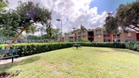 This single-floor residence is situated within a vibrant multi-building community that showcases a warm, tropical aesthetic characteristic of Florida living. The exterior grounds are a standout feature, with expansive, well-manicured lawns framed by neatly trimmed hedgerows and mature palm and shade trees that lend the property a lush, park-like atmosphere. A picnic area with seating is nestled within the green space, offering a comfortable outdoor gathering spot for residents. The community grounds are complemented by tennis or sport courts visible in the background, enclosed by chain-link fencing, and illuminated by tall light poles for evening use. The surrounding residential buildings display a colorful palette of warm reds and tans with covered balconies, contributing to a cohesive and inviting architectural style. The open sky above allows abundant natural light to fill the outdoor spaces throughout the day. While the interior images available are limited in visibility, the single-story layout of the unit suggests an accessible and efficiently designed floor plan. Overall, the property benefits greatly from its well-kept communal outdoor environment, offering residents a blend of green space, recreational facilities, and a relaxed, sun-drenched community setting.
