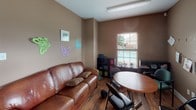 This single-story facility offers a warm, residential atmosphere designed to foster comfort and community. The space is finished throughout with rich hardwood flooring and neutral taupe-toned walls that create a cohesive, calming environment.

The main gathering area is particularly striking, boasting soaring ceilings that amplify the sense of openness. Multiple windows dressed with decorative valances and white horizontal blinds allow generous natural light to fill the room, while half-moon sconce lighting accents the walls. A prominent decorative entry door with leaded glass sidelights serves as a welcoming focal point, accompanied by an adjacent exit door. Round wooden tables with black folding chairs are arranged throughout this area to accommodate group activities or meetings. A stone-accented fireplace or feature wall is visible from the main space, adding architectural character.

A secondary lounge area provides a more intimate setting, furnished with brown leather sofas, a club chair, and a coffee table, creating a comfortable space for smaller gatherings or one-on-one interactions. Decorative elements such as colorful butterfly wall art and framed prints give this room a lighter, more playful feel. An additional room with leather seating, a patterned area rug, and curtained walls offers a quieter, more private setting. Together, the rooms reflect a thoughtfully designed facility built for both community engagement and individual comfort.