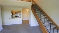 This two-story residence offers a well-organized main level that integrates the living and kitchen areas into a cohesive, open layout. The living space is finished with wall-to-wall carpeting in a neutral tone, lending a comfortable, understated feel throughout. A ceiling fan overhead provides both light and ventilation to the main living area.

The kitchen is positioned adjacent to the living space and is accessible via a breakfast bar-style counter with granite or stone-look countertops. Warm wood cabinetry lines the kitchen walls, complemented by stainless steel appliances including a microwave and a gas range, offering a functional and tidy cooking environment. The kitchen pass-through design maintains a visual connection to the rest of the main floor.

A staircase positioned along one wall leads to the upper level, featuring a wood handrail and metal baluster railing system that adds a contemporary touch to the interior. The upper landing is visible from below, with matching metal railings creating a consistent design element across both floors. Neutral wall tones throughout contribute to a clean, move-in-ready aesthetic.